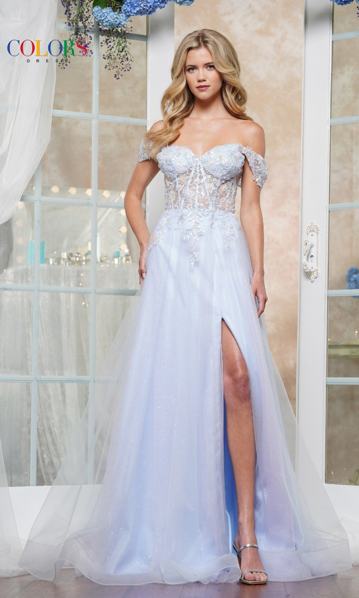 A woman indoors stands in front of glass doors wearing the Off-Shoulder Glitter Tulle Prom Ball Gown: Colors Dress 3498, featuring a light blue hue, corset bodice, lace details, and a thigh-high slit.